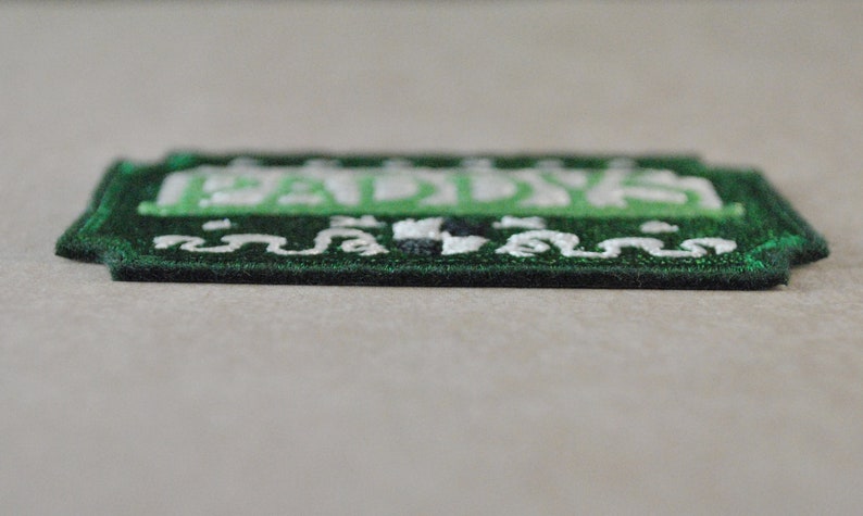 May include: Green embroidered patch with white lettering that reads "Paddy's". The patch has a decorative border.