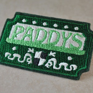 May include: Green and white embroidered patch with the word "Paddy's" in white lettering. The patch has a green border and a white shamrock design.