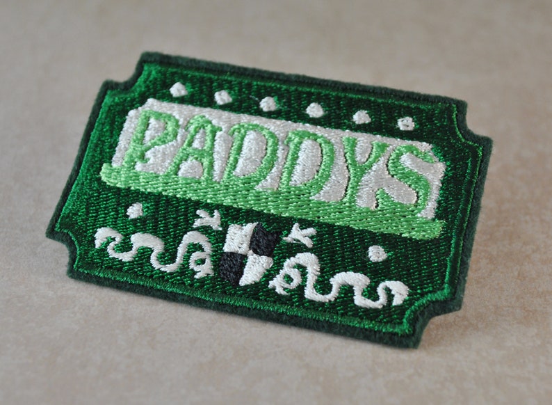 May include: Green and white embroidered patch with the word "Paddy's" in white lettering. The patch has a green border and a white shamrock design.