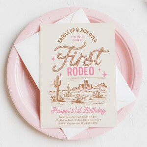 May include: A pink and white invitation with a brown illustration of a desert landscape. The text reads "Saddle Up & Ride Over It's Our Girl's First Rodeo" and "Harper's 1st Birthday".