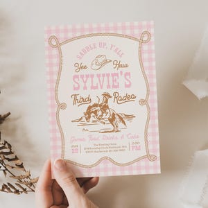 May include: A rectangular invitation card with a pink and white gingham border. The center features brown text reading SADDLE UP, Y'ALL, SYLVIE'S THIRD RODEO, and Yee Haw, with a brown illustration of a person riding a bucking horse and a cowboy hat.