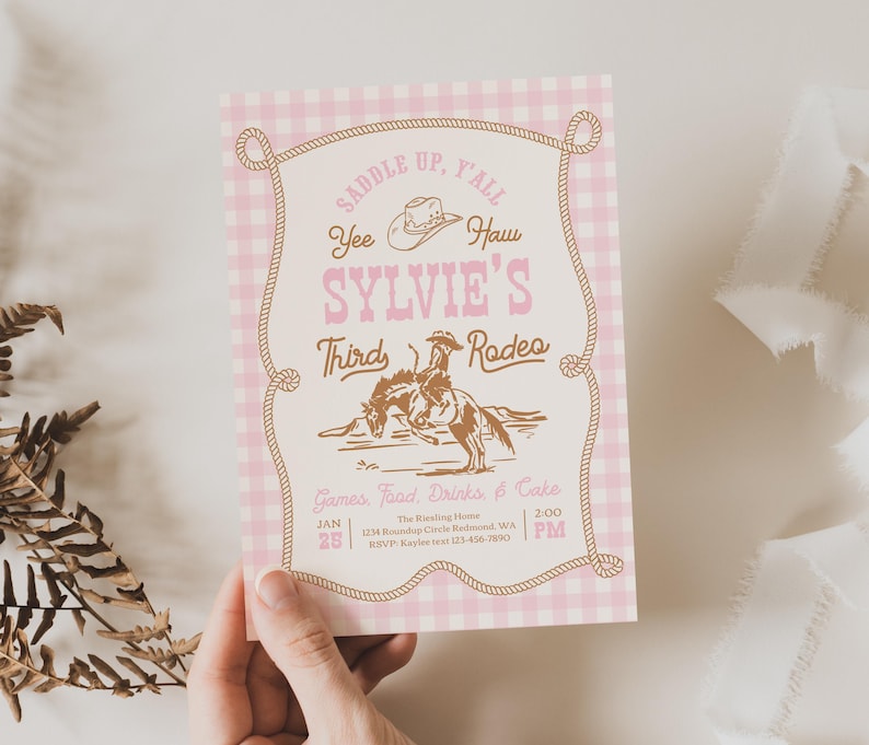 May include: A rectangular invitation card with a pink and white gingham border. The center features brown text reading SADDLE UP, Y'ALL, SYLVIE'S THIRD RODEO, and Yee Haw, with a brown illustration of a person riding a bucking horse and a cowboy hat.