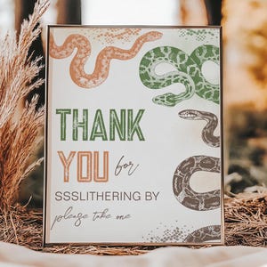 Könnte beinhalten: Ein goldfarbener Rahmen mit einem weißen Hintergrund und darauf gedruckten grünen, braunen und orangefarbenen Schlangen. Das Schild sagt "THANK YOU for SSSLLITHERIN BY please take one".
