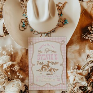 May include: A rectangular invitation card with a light pink gingham border and gold rope frame. The text reads: Saddle Up, Y'all, Yee Haw, Sylvie's Third Rodeo, Games Food Drinks & Cake, Jan 25, The Riving Home, 1234 Roundup Circle, RSVP: Kaylee.