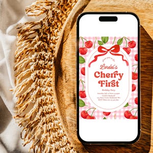 May include: A smartphone displays a birthday party invitation with a cherry theme. The invitation features red cherries, green leaves, and the text "Lorelei's Cherry First" on a pink gingham background. The phone rests on a wooden tray.