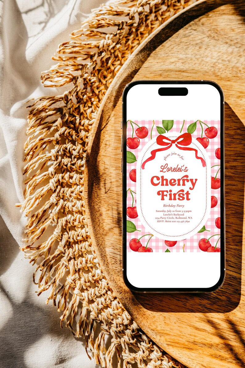 May include: A smartphone displays a birthday party invitation with a cherry theme. The invitation features red cherries, green leaves, and the text "Lorelei's Cherry First" on a pink gingham background. The phone rests on a wooden tray.