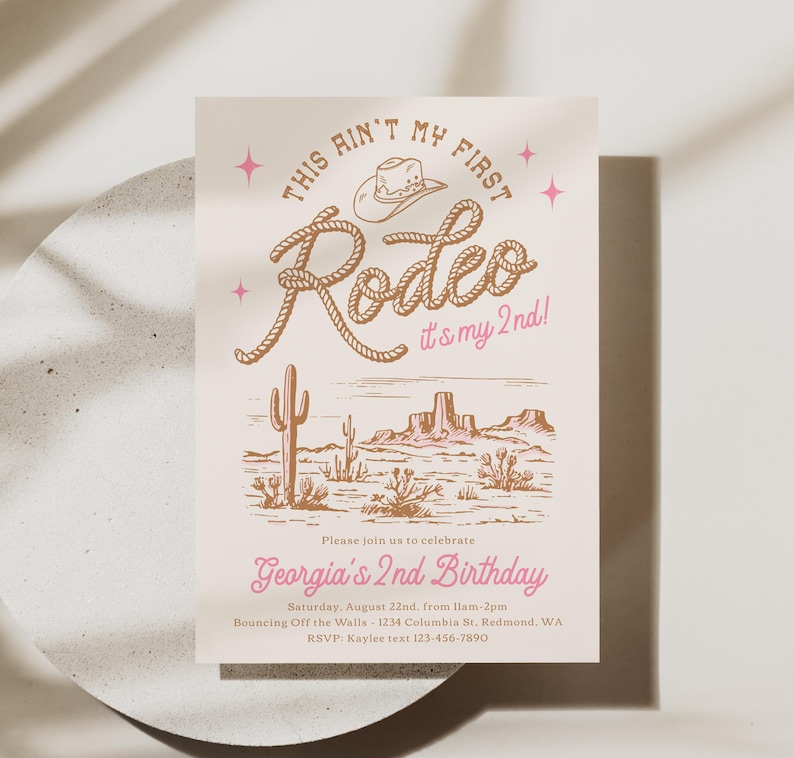 May include: A rectangular birthday invitation card on a light cream background featuring a brown cowboy hat and rope text that reads This Ain't My First Rodeo it's my 2nd! with pink stars and text Georgia's 2nd Birthday and event details.