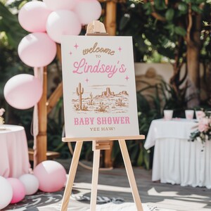 May include: A pink and gold welcome sign with a desert scene and the text "Welcome to Lindsey's Baby Shower Yee Haw!" on a wooden easel.