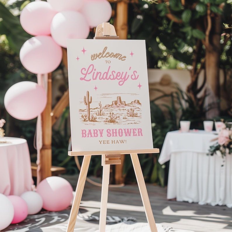 May include: A pink and gold welcome sign with a desert scene and the text "Welcome to Lindsey's Baby Shower Yee Haw!" on a wooden easel.
