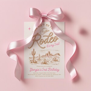 May include: A rectangular invitation card with a pink satin bow at the top. The card features a brown western-themed illustration of a cowboy hat, rope-style text reading "This ain't my first rodeo, it's my 2nd!", and a desert landscape with cacti.