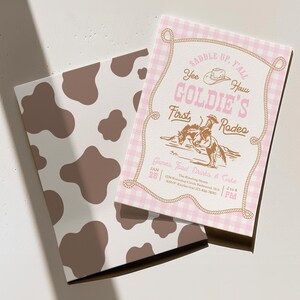 May include: A rectangular invitation card with a pink and white gingham border and a brown rope frame. The text reads SADDLE UP, Y'ALL, YEE HAW, GOLDIE'S FIRST RODEO, and GAMES, FOOD, DRINKS & CAKE. A brown cow print card is positioned underneath.