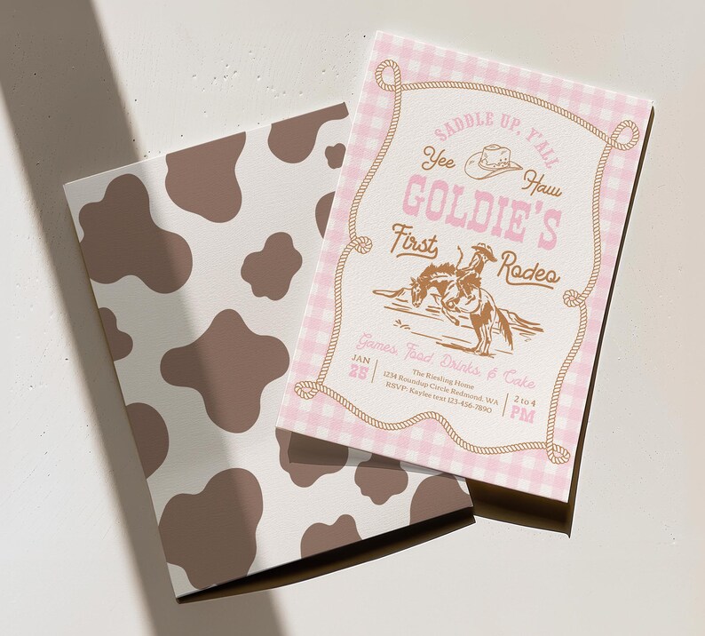 May include: A rectangular invitation card with a pink and white gingham border and a brown rope frame. The text reads SADDLE UP, Y'ALL, YEE HAW, GOLDIE'S FIRST RODEO, and GAMES, FOOD, DRINKS & CAKE. A brown cow print card is positioned underneath.