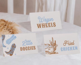 May include: Three white cards with brown text and illustrations. The first card says "Wagon Wheels" in blue and brown text. The second card says "Little Doggies" in blue and brown text and has a blue illustration of cowboy boots. The third card says "Fried Chicken" in blue and brown text and has a brown illustration of a cactus.