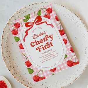 May include: A pink gingham patterned invitation with red cherries and green leaves. The invitation reads "Lorelei's Cherry First" with event details. The invitation is on a white plate.