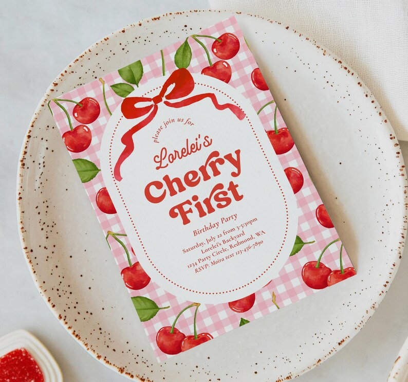 May include: A pink gingham patterned invitation with red cherries and green leaves. The invitation reads "Lorelei's Cherry First" with event details. The invitation is on a white plate.