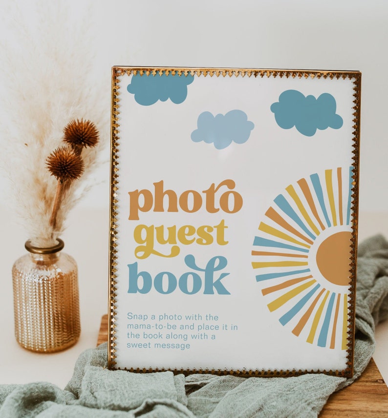 K&ouml;nnte beinhalten: Ein goldfarbener Rahmen mit wei&szlig;em Hintergrund und dem Text "photo guest book" in einer Retro-Schrift. Das Schild zeigt au&szlig;erdem eine stilisierte Sonne und Wolken in Gelb, Blau und Orange.