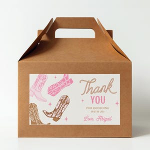 Puede incluir: Una caja de cartón marrón con asa. La caja tiene una etiqueta blanca con botas de vaquero rosas y el texto "Gracias por el rodeo con nosotros! Con cariño, Abigail".