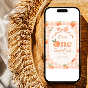 May include: A smartphone displays a peach-themed invitation for a first birthday. The invitation features a peach and bow design with the text "Eloise is One Sweet Peach" on a gingham background. The phone rests on a wooden tray.