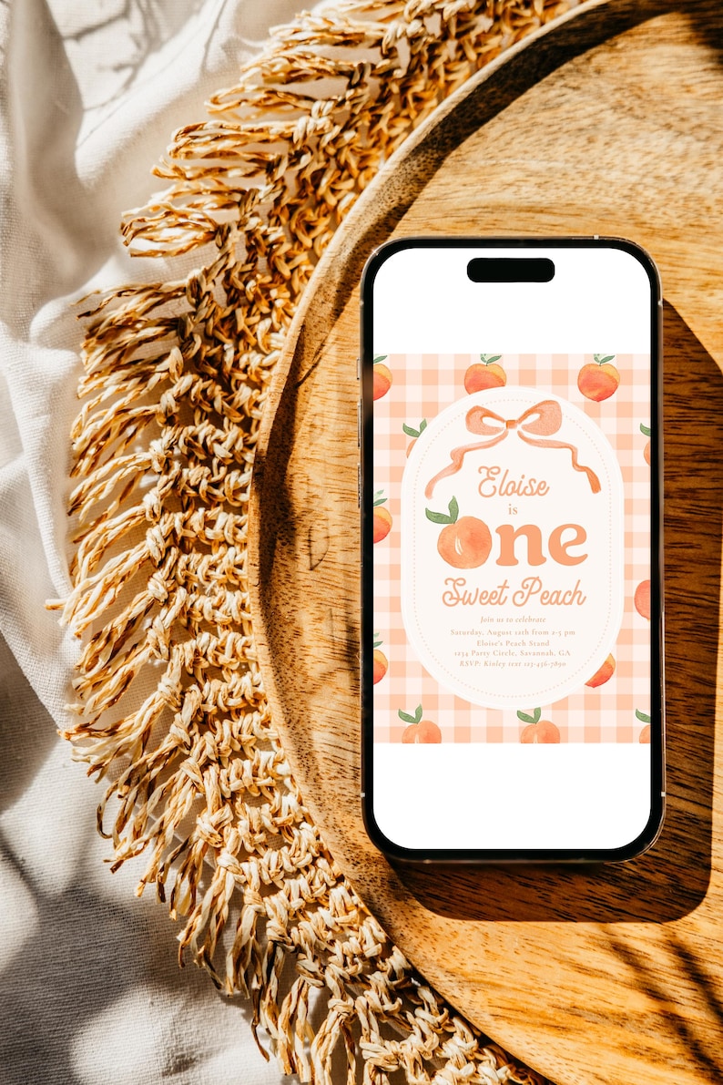 May include: A smartphone displays a peach-themed invitation for a first birthday. The invitation features a peach and bow design with the text "Eloise is One Sweet Peach" on a gingham background. The phone rests on a wooden tray.