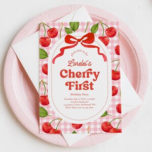 May include: A pink and white gingham patterned invitation with red cherries and green leaves. The invitation reads "Lorelei's Cherry First Birthday Party" with event details. The invitation is on a pink plate with a white envelope.