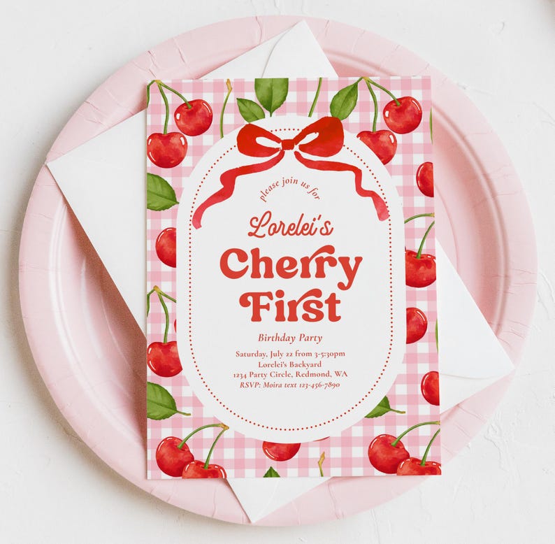 May include: A pink and white gingham patterned invitation with red cherries and green leaves. The invitation reads "Lorelei's Cherry First Birthday Party" with event details. The invitation is on a pink plate with a white envelope.