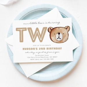 May include: A birthday invitation featuring a watercolor illustration of a teddy bear's face. The invitation reads "our little bear is turning TWO" in brown letters. The text also includes the name "Hudson" and party details.