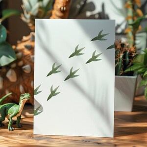 May include: A white card with green dinosaur footprints printed on it. The card is sitting on a wooden table with a green dinosaur toy in the foreground.