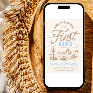 May include: A smartphone displaying a digital invitation for a first birthday party with a western theme. The invitation features a drawing of a desert landscape with cacti and mountains, and the text "Saddle Up & Ride Over It's Our Boy's First Rodeo". The invitation also includes the birthday boy's name, date, time, location, and RSVP information.