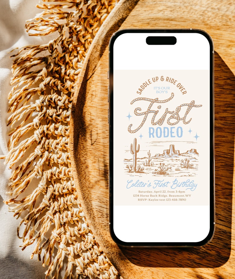 May include: A smartphone displaying a digital invitation for a first birthday party with a western theme. The invitation features a drawing of a desert landscape with cacti and mountains, and the text "Saddle Up & Ride Over It's Our Boy's First Rodeo". The invitation also includes the birthday boy's name, date, time, location, and RSVP information.
