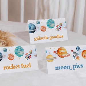 May include: Three space-themed table cards with watercolour illustrations of planets and rockets. The cards read "galactic goodies", "rocket fuel", and "moon pies".