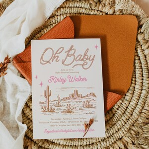 May include: A pink and brown baby shower invitation with a western theme. The invitation says "Oh Baby" in a pink rope design and "Kinley Walker" in pink lettering. The invitation also includes a desert landscape illustration with cacti and mountains.