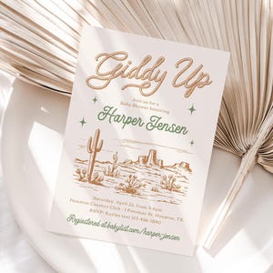 May include: A baby shower invitation with a western theme. The invitation features a drawing of a desert landscape with cacti and mountains. The text on the invitation reads "Giddy Up" and "Join us for a Baby Shower honouring Harper Jensen".