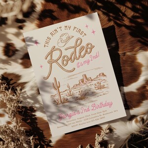 May include: A rectangular invitation card with a cream background featuring the text "THIS AIN'T MY FIRST Rodeo it's my 2nd!" in brown and pink. Below is a desert illustration with cacti and mesas, followed by "Georgia's 2nd Birthday" and event details.