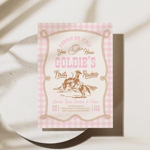 May include: A rectangular invitation card with a light pink and white gingham border. The center features a brown illustration of a person riding a bucking horse, a cowboy hat, and text reading: SADDLE UP, Y'ALL, Yee Haw, GOLDIE'S First Rodeo, Games, Food, Drinks & Cake.
