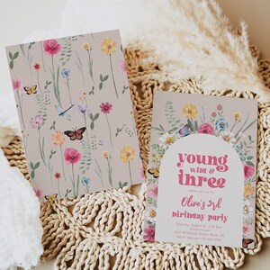 May include: A floral-themed birthday party invitation with a pink and white color scheme. The invitation features a whimsical illustration of flowers, butterflies, and a dragonfly. The text on the invitation reads "young wild & three" and "Olive's 3rd birthday party".