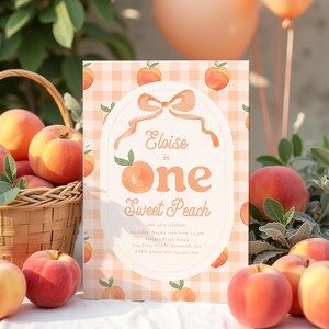 May include: A peach-themed invitation with the text "Eloise is One Sweet Peach" is surrounded by fresh peaches and a wicker basket. The invitation has a peach gingham background and a peach-colored bow.