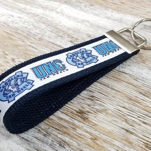 May include: A blue and white UNC Tar Heels keychain with a black strap and a silver key ring. The keychain features the UNC logo and the words "UNC Tar Heels".