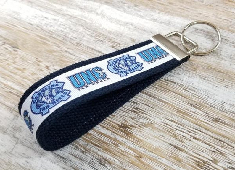 May include: A blue and white UNC Tar Heels keychain with a black strap and a silver key ring. The keychain features the UNC logo and the words "UNC Tar Heels".