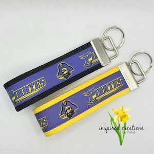 May include: Two keychains with a purple and yellow design. The keychains have a black and yellow background with the word "PIRATES" in gold lettering. Each keychain features a pirate logo and a silver key ring.