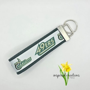 May include: A key fob with a white ribbon featuring the text "49ERS" in green and gold, and "UNC CHARLOTTE" in green. The fob has a silver-colored metal clasp and a key ring. The background is white.