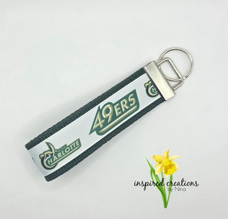 May include: A key fob with a white ribbon featuring the text "49ERS" in green and gold, and "UNC CHARLOTTE" in green. The fob has a silver-colored metal clasp and a key ring. The background is white.