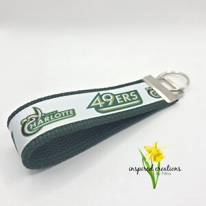 May include: A green and white keychain with the text "49ERS" and "UNC CHARLOTTE". The keychain has a silver metal ring and a green fabric loop. A yellow flower is in the bottom right corner.