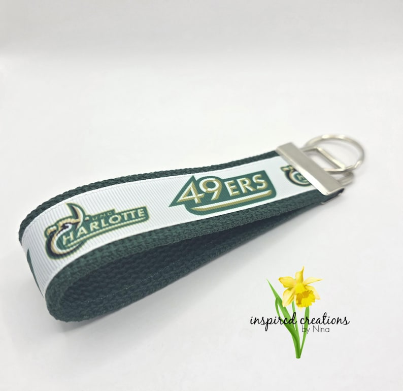 May include: A green and white keychain with the text "49ERS" and "UNC CHARLOTTE". The keychain has a silver metal ring and a green fabric loop. A yellow flower is in the bottom right corner.