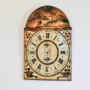 Puede incluir: Una esfera de reloj vintage con números romanos y una escena pintada de una pareja caminando en un jardín. La esfera del reloj es blanca con números romanos negros y un borde marrón. La escena está pintada en colores apagados e incluye una pequeña casa y árboles.