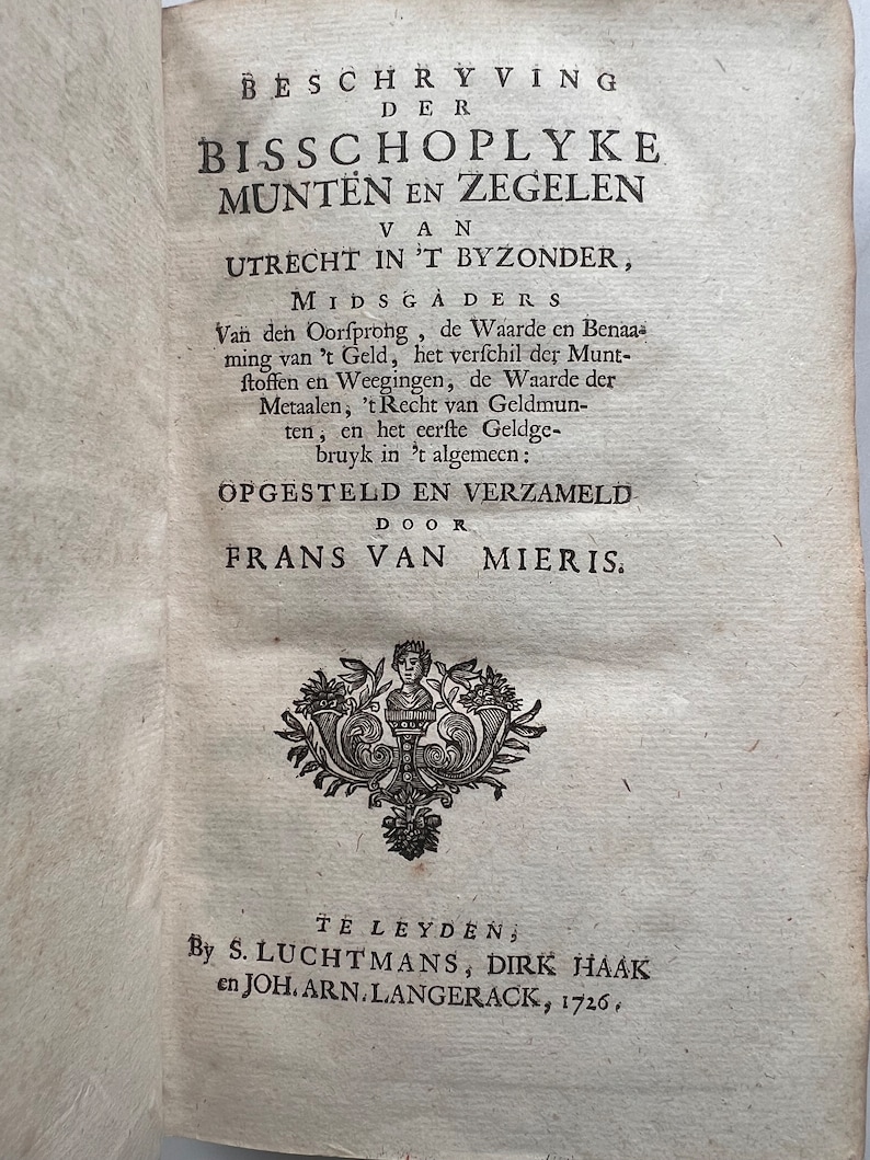 May include: A black and white image of a book cover with the title "Beschryving der Bisschoplyke Munten en Zegelen van Utrecht in 't Byzonder, Midsgaders" and the author "Frans van Mieris".
