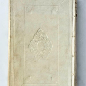 May include: The cover of an antique book with a decorative design in the center. The cover is made of a cream-colored material and has a worn appearance.