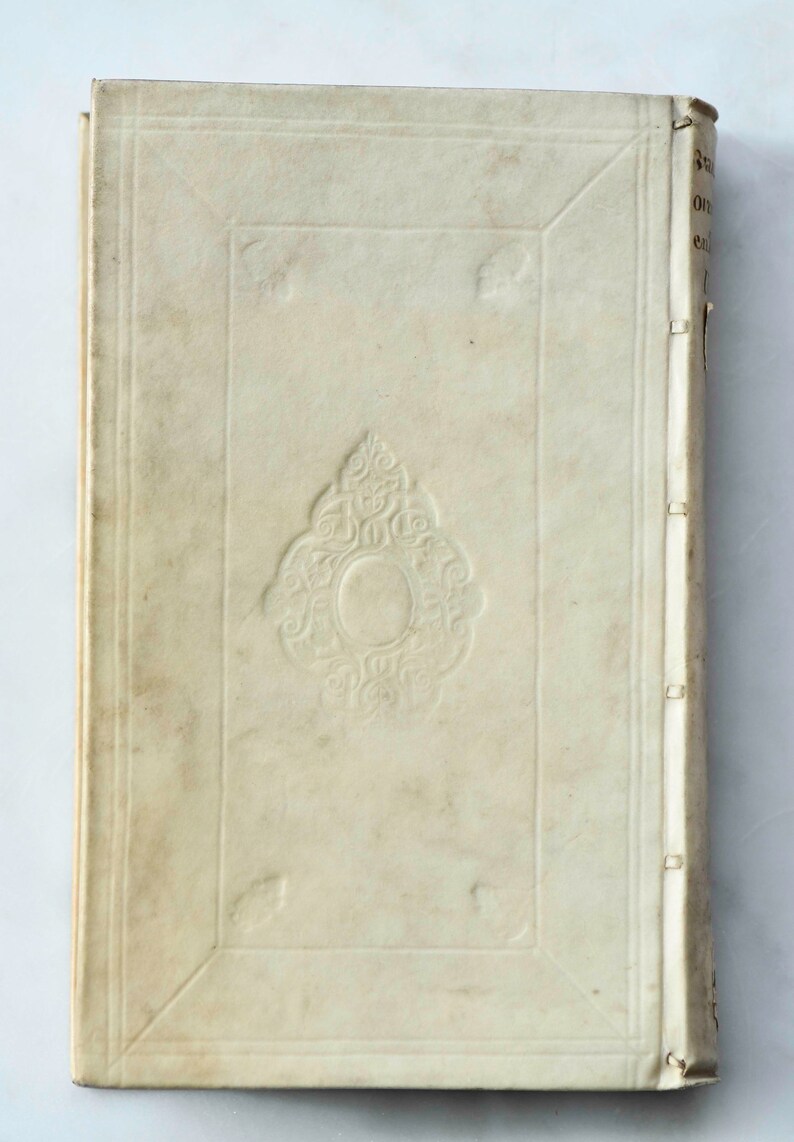 May include: The cover of an antique book with a decorative design in the center. The cover is made of a cream-colored material and has a worn appearance.