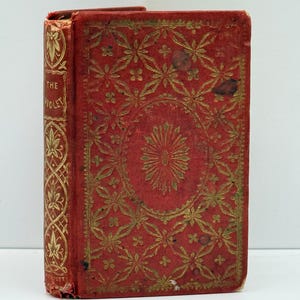 May include: A red hardcover book with gold embossed floral and geometric patterns on the front cover and spine. The spine features the text THE VIOLET in gold lettering. The cover shows signs of wear with darkened spots and frayed edges.