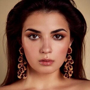 May include: A woman with long dark hair wearing a brown and white animal print off-the-shoulder top and large gold and burgundy beaded earrings.