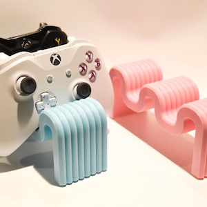 May include: A white and black video game controller is resting on a light blue and pink stand. The stand is made of plastic and has a wavy design.